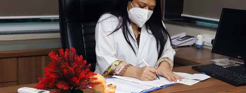 Dr. Iram seated in her office, wearing a mask and white coat, while reviewing patient documents.